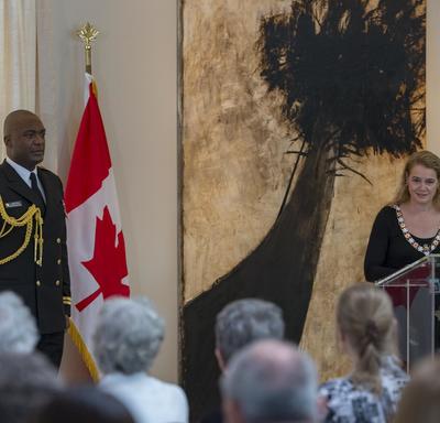 The Governor General stands behind a podium and faces a room full of people.  She delivers a speech. 