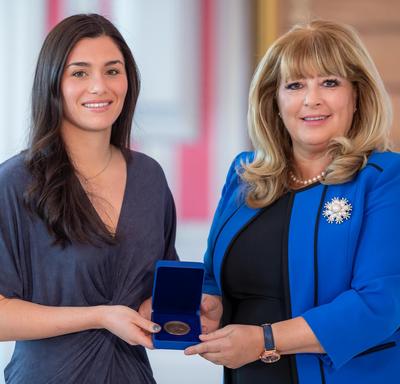 On the left, a female dark hair university student is holding an opened blue box containing a medal. A blond woman wearing a blue jacket is on the right.