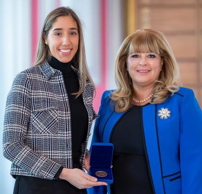 On the left, a tall female university student is holding an opened blue box containing a medal. A blond woman wearing a blue jacket is on the right.