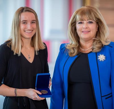 On the left, a blond female university student is holding an opened blue box containing a medal. A blond woman wearing a blue jacket is on the right.