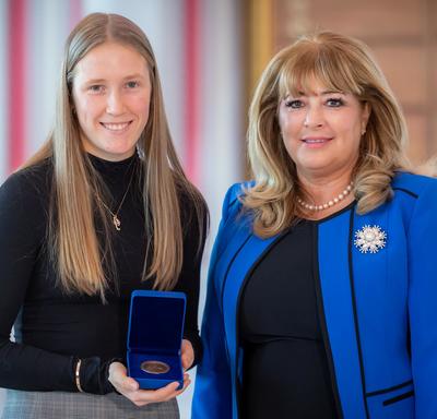  gauche, une étudiante universitaire blonde tient une boîte bleue ouverte contenant une médaille. Une femme blonde portant une veste bleue se trouve à droite.