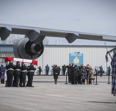 CAF members carry a coffin to the hearse. A bagpiper is in the forefront of the picture.