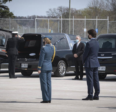 The Governor General salutes a coffin as it is placed in a hearse. The Prime Minister is standing behind her. 