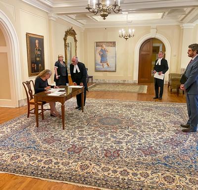 Vue de côté. Assise sur une chaise de bois, à une table de bois, la gouverneure générale signe un document.