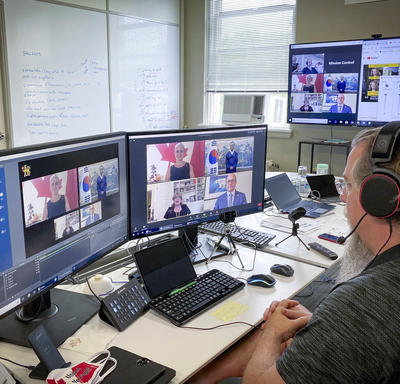 A man with a head set is sitting at a computer, looking at two large screens in front of him. A virtual conversation with 4 participants seems to be taking place.