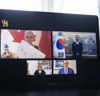 A computer screen split in 4 rectangles with a person in each one, 2 women and 2 men. Top left rectangle shows a Canada flag behind a woman, holding a certificate-like paper. On her right, a man is standing next to the Korea flag.