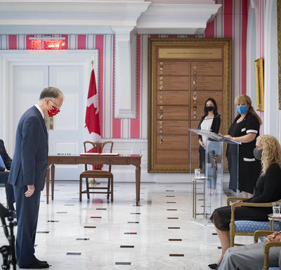 A man in a suit is standing in front of the Governor General who is seated. He is acknowledging her with a nod. The Prime Minister looks on from the side.