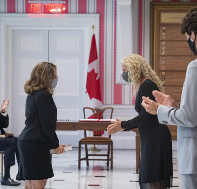 A woman is standing facing Governor General Payette and Prime Minister Trudeau who are applauding.
