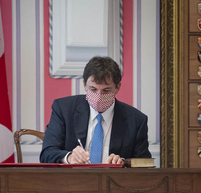 A man is seated at a desk to the right of a large Canadian flag. He is signing a document.