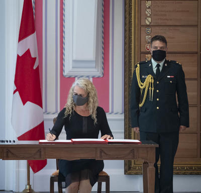 A woman is seated at a desk between a large Canadian flag and a soldier in dress uniform. She is signing a document.