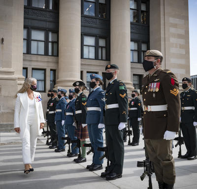 A woman dressed in white walks past several military members who are standing at attention. Some are wearing green, others are dressed in black. There is also a man in uniform to the woman’s right. All are wearing masks.