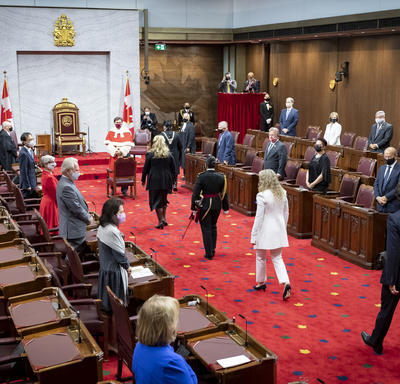 A procession of people are walking along a red carpet towards the front of the room. Three of the individuals are wearing black. One woman is wearing white. There is a throne chair flanked by two Canada flags. 