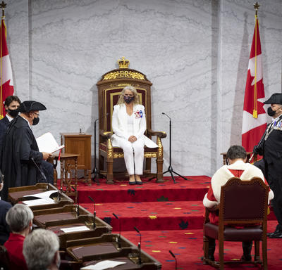A woman dressed in a white suit is sitting on a throne chair. There are Canada flags on either side of the platform. There is a man sitting in a chair facing her. He is wearing a red and white cape.
