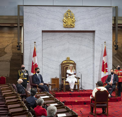 Une femme en costume blanc est assise sur un trône et tient un porte-document noir. Un homme est assis face à elle sur un tapis rouge. On peut également voir d'autres personnes dans le public, assises.