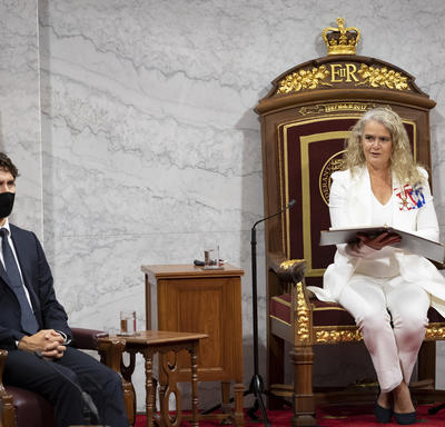 A woman dressed in a white suit is sitting in a large throne chair holding a black binder. There is a small wooden table to her right. There is also a man to her right. He is dressed in a black suit with a navy blue tie.