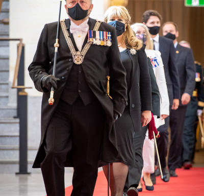 Une file de personnes marchent vers la caméra sur un tapis rouge. L'homme qui dirige le groupe porte un uniforme noir décoré, avec un bâton noir et doré. Il porte également un chapeau noir et un masque noir.