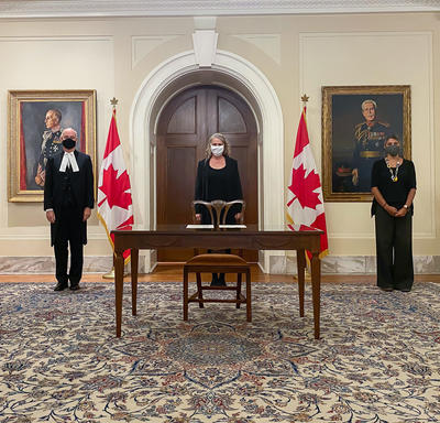 Quatre personnes portant des masques faciaux se tiennent au fond de la salle. Il y a une table et une chaise en bois devant la femme au centre de la photo. Il y a deux drapeaux canadiens de chaque côté d'elle.
