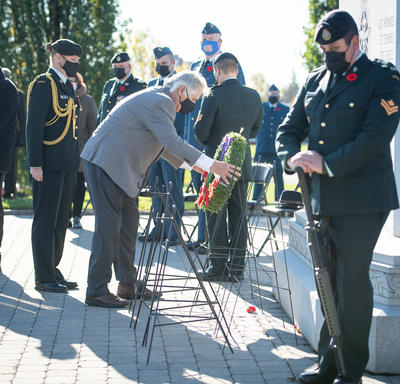 Mr. Whit Grant Fraser is laying a wreath in front of a stone memorial. Several members of the Canadian Armed Forces are standing around him, wearing masks. Behind him, there is a man in a black coat standing at a podium. They are outside and it is sunny.