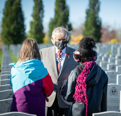 Mr. Whit Grant Fraser is standing with his back to many rows of headstones. He is wearing a mask and is talking to 2 students.