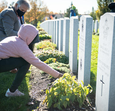 A student is kneeling in front of a headstone and is placing a poppy at its base. Mr. Whit Grant Fraser is crouching next to her, looking at the poppy. He is wearing a mask. There are many rows of headstones in the background.