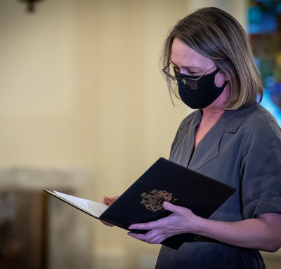 A woman reading from a document. She is wearing a black mask.