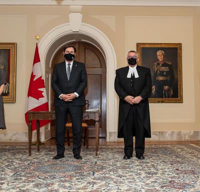 Quatre personnes, dont l'administrateur et le secrétaire, sont debout, tournées vers l'avant. Toutes portent des masques. Un drapeau canadien en arrière-plan. 