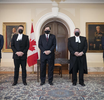 L’administrateur se tient entre quatre personnes. Toutes les personnes portent des masques. Un drapeax du Canada se trouve à l’arrière-plan.