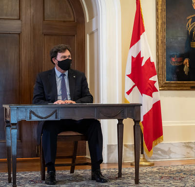 L’administrateur est assis à une table. Le secrétaire se tient à sa gauche. On aperçoit un grand drapeau du Canada derrière et entre les deux hommes.