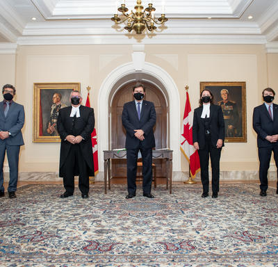 L’administrateur se tient entre quatre personnes. Toutes les personnes portent des masques. Deux drapeaux du Canada se trouvent à l’arrière-plan.