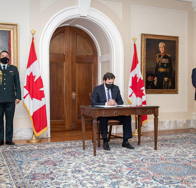 The Administrator sitting at a table. The Secretary is standing to his left while an aide-de-camp stands to his right.