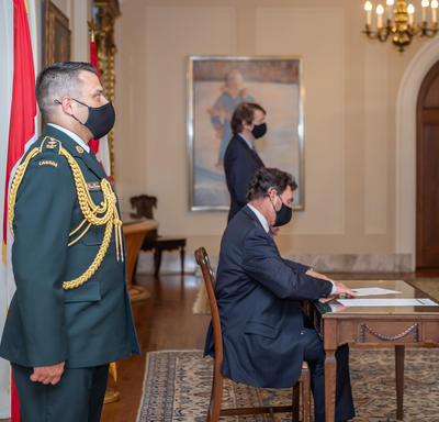 An aide-de-camp standing as the Administrator sits at a table. The Secretary is visible in the background.