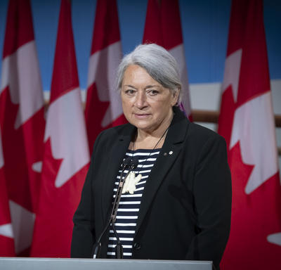 La gouverneure générale désignée Mary May Simon se tient face à un podium devant plusieurs drapeaux du Canada.