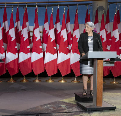 Le premier ministre Justin Trudeau et la gouverneure générale désignée Mary May Simon se tiennent chacun face à un podium devant plusieurs drapeaux du Canada derrière eux.