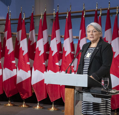 Le premier ministre Justin Trudeau et la gouverneure générale désignée Mary May May Simon se tiennent chacun face à un podium devant plusieurs drapeaux du Canada derrière eux. Mary Simon parle. Justin Trudeau la regarde.