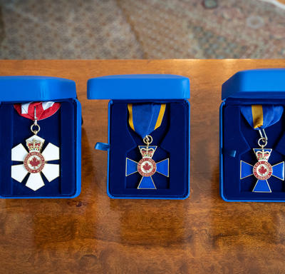 Three Canadian order decorations are positioned on a wood table in individual blue suede boxes. The first decoration is red and white with a red and white ribbon. The second and third decorations are blue with blue ribbon with a yellow border. 