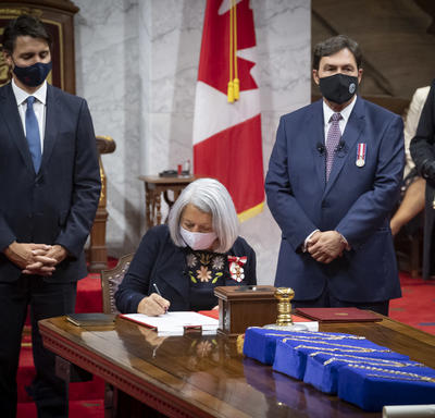 Mary May Simon signing a document in the Senate. The Prime Minister and the Supreme Court Chief Justice Richard Wagner stand on either side of her. 