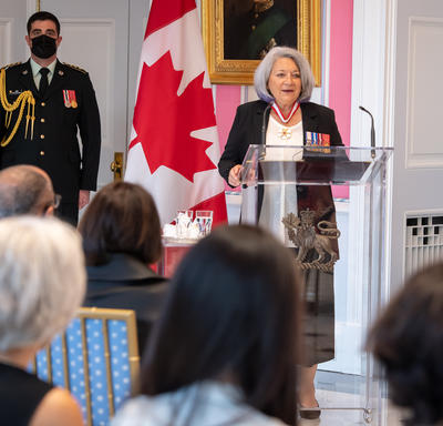 Governor General Mary May Simon stands at a glass podium addressing the audience in the Tent Room at Rideau Hall. There is a large Canadian flag behind her.
