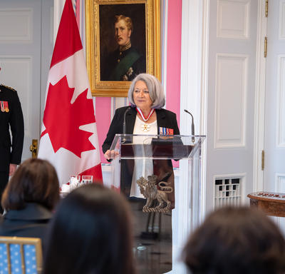 La gouverneure générale Mary May Simon, debout derrière un lutrin en verre, s’adresse au public rassemblé dans la salle de la tente de Rideau Hall. Un imposant drapeau canadien décore le fond de la pièce.