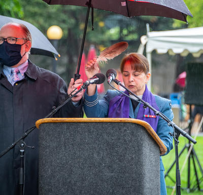A woman stands at a podium. A man holds an umbrella over her to protect her against the rain.