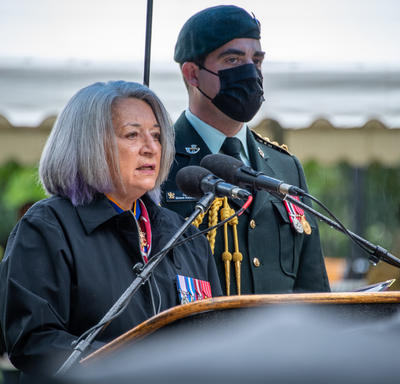 La gouverneure générale Mary May Simon prononce son allocution. Elle est dehors. Un homme en uniforme militaire portant un masque se tient derrière elle. Il pleut. 