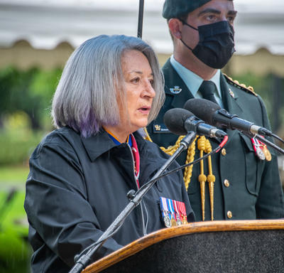 La gouverneure générale prononce son allocution. Un homme en uniforme militaire portant un masque se tient derrière elle. Il pleut. 
