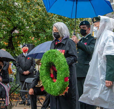 The Governor General lays a wreath at the base of the National Aboriginal Veterans Monument. Feathers are visible on the left side of the photo. In the background are trees and a small crowd of people wearing masks.