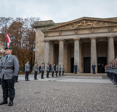 Grand bâtiment à l’arrière-plan. Des membres de l’armée allemande sont debout, en uniforme. Ils sont à l’extérieur.