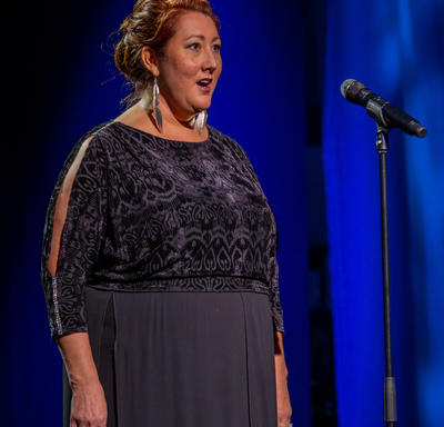 A lady is singing. She is standing at a microphone. She is dressed in formal evening wear. 