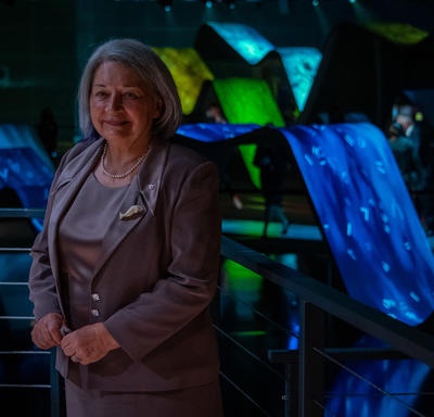 Governor General Mary May Simon is standing in front of a large illuminated art display made of blue, green, and yellow ramps with projections on them. She is smiling.