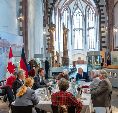 The Governor General and Mr. Fraser are sitting at a table with six other people. In the background there is a Canadian flag and to its left a German flag. There is also a suit of armour on a display rack.