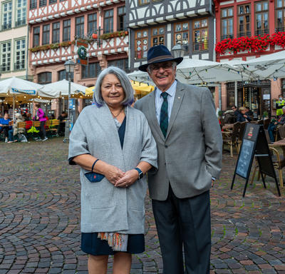 Governor General Mary May Simon and Mr. Fraser pose on a street in Old Town Frankfurt. Mr. Fraser has his arm around Ms. May Simon and they are smiling. There are colourful buildings in the background and several vendor tents.