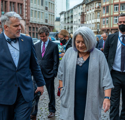 Governor General Mary May Simon on a street in Old Town Frankfurt. She is beside a man who is accompanying her. There are colourful buildings in the background and several vendor tents.uildings in the background and several vendor tents.