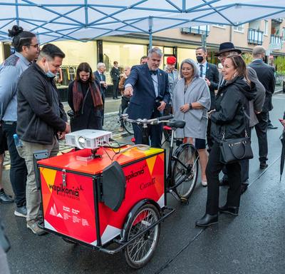 Their Excellencies are outdoors. They are standing with a group of people admiring a bicycle cart from the Frankfurt Book Fair. 