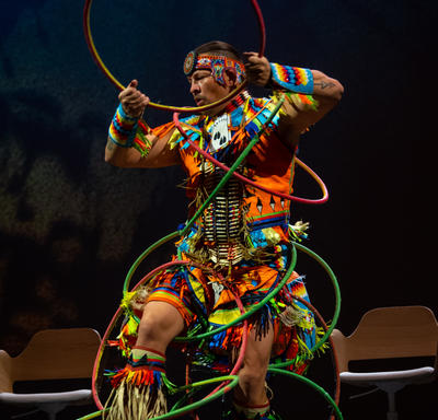 Dallas Arcand est une scène, il exécute une danse autochtone avec 11 cerceaux. Il porte une tenue traditionnelle très colorée. Derrière lui se trouvent plusieurs chaises en bois.